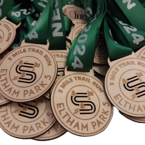 A pile of sustainable wooden medals made in the UK with green ribbons, each engraved with 5 Mile Trail Run, Eltham Park 5, 2024, and a stylised running shoe design.