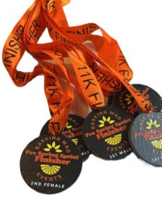 A pile of black medals with orange ribbons labelled 11K FINISHER and Barking Mad Events. The medals read “Pre Spring Sprint Finisher” and indicate placings like “2nd Female” and “1st Male”.