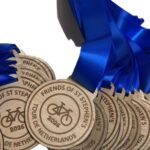 A pile of wooden medals with blue ribbons, engraved with a bicycle and the words FRIENDS OF ST STEPHEN'S TOUR DE NETHERLANDS 2026.