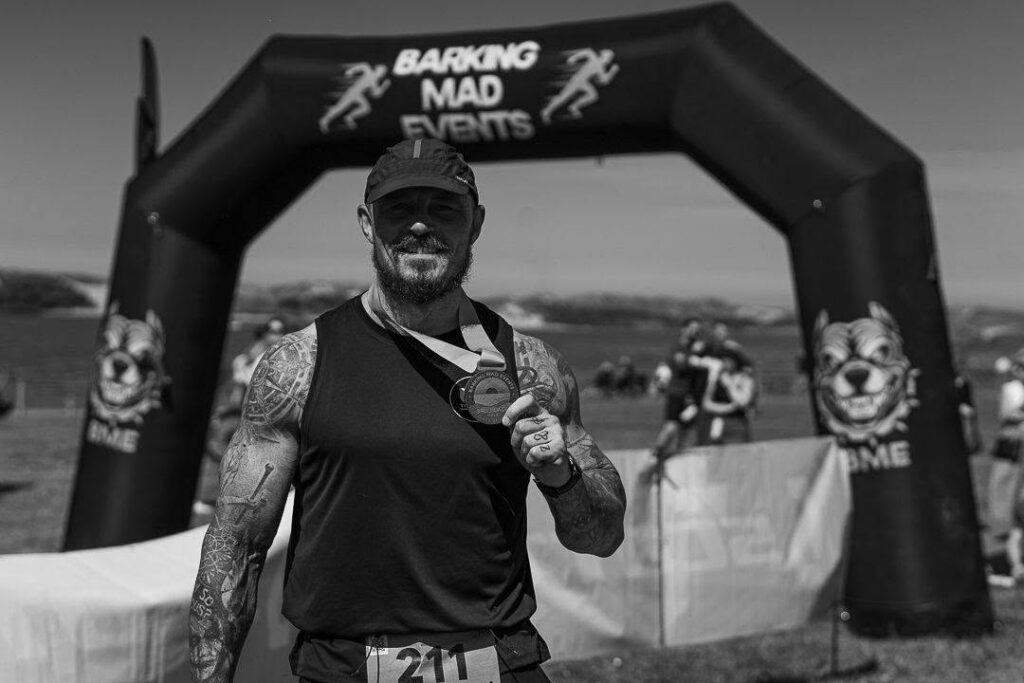 A man with tattoos holds a medal and smiles in front of an inflatable arch that reads Barking Mad Events. Other participants can be seen in the background at an outdoor event.