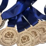 Several wooden medals with blue ribbons, engraved with BORDERS TOWN CONFERENCE WINNERS 25/26 BOYS U15, are laid out overlapping each other on a white background.