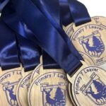 Wooden medals with blue ribbons, engraved with a stag, trees, Primary 7 Leaver, and the year 2023, displayed in a pile.