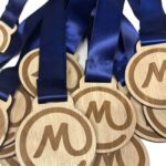 A pile of wooden medals with an engraved M and blue satin ribbons attached, arranged overlapping each other on a white background.