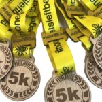 Several wooden 5k medals with bright yellow ribbons that say #nooneleftbehind and #MilesShyClub are arranged in a pile.