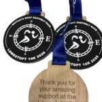 Three medals with blue ribbons. Two black medals show a running figure, compass, and text Lowestoft 10K 2026. The back of a wooden medal reads: Thank you for your amazing support at the Lowestoft 10K 2026.