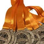 A row of wooden sports medals with orange satin ribbons, engraved with ARAN KAKAS SPORTS DAY and a trophy illustration.
