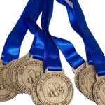 Seven wooden medals with blue ribbons read Busy Bees Nursery Ashton Leavers 2023 and feature a cartoon bee design. The medals are stacked and arranged diagonally against a white background.