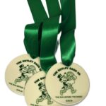 Three white medals with green ribbons for The Butley Dash 5K: The Run Before the Rings event, featuring cartoon runners and the date 12.06.26.