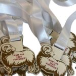A pile of white ribbons attached to ram-shaped medals that read RAM 100 2026 FINISHER in red letters.