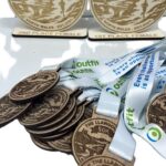 Wooden medals and trophies for the Llangollen 50K race in North Wales are shown. The medals have event engravings, and the trophies are labelled 1st Place Female and 2nd Place Female.
