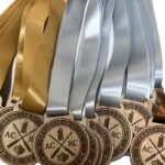 A row of gold and silver medals with light-coloured ribbons, engraved with ACRC Ardglass Coastal Rowing Club and an oar design, displayed against a white background.