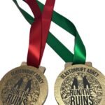 Two gold medals with red and green ribbons read Glastonbury Abbey Run the Ruins. Both feature artwork of ruins and intricate designs.