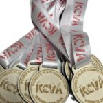 A pile of wooden medals with Women’s Plate Competition Winner 2025–2026 KEYA engraved, attached to white ribbons with red writing.