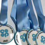 Four silver medals with blue ribbons labelled CO-OP CHARITY GOLF DAY 2023 2ND are arranged side by side on a white surface.