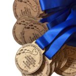A pile of circular wooden commemorative medals, each with blue ribbons and engraved text that reads: Walking every step for a cause, 27 miles completed in memory of Nick McGourty, 11th September 2026.