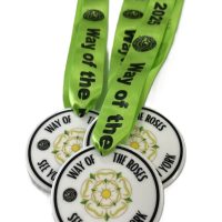 Three white Eco Medals with a yellow and green rose design and the words WAY OF THE ROSES SEE YORK RUN YORK are attached to bright green ribbons that read Way of the Roses 2025 in black text.