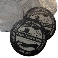 Round coasters with a silver and black design promoting Royal Deeside 2025: Six Days of Scottish Orienteering, featuring a castle, trees, and orienteers, along with event dates and surrounding location names.