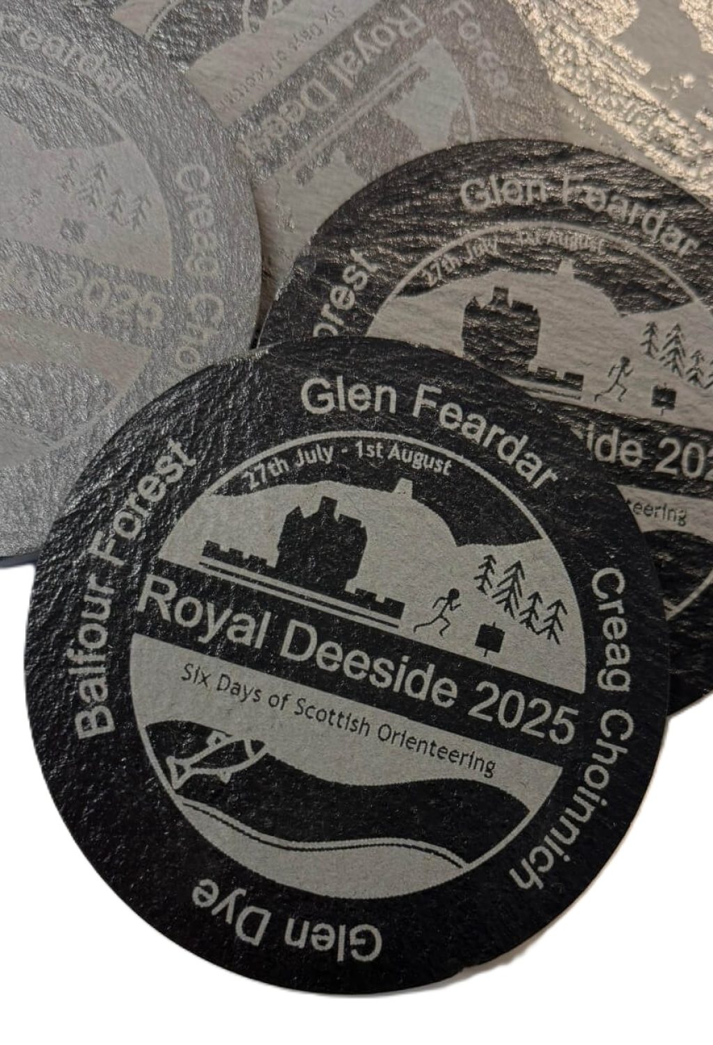 Round coasters with a silver and black design promoting Royal Deeside 2025: Six Days of Scottish Orienteering, featuring a castle, trees, and orienteers, along with event dates and surrounding location names.
