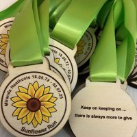 A stack of wooden Sunflower Run medals with green ribbons. The medal front shows a sunflower and event details, whilst the back reads, Keep on keeping on... there is always more to give.