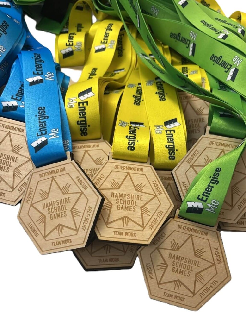 A pile of wooden hexagonal medals with blue, yellow, and green ribbons labelled Energise Me. The medals are engraved with Hampshire School Games, DETERMINATION, and TEAM WORK.