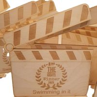 A wooden film clapperboard trophy, part of the Eco Medals collection, is engraved with “THE SHORTEST NIGHTS Winner 2025,” “Swimming in it,” and “BEST DARK COMEDY.” Several similar trophies are visible in the background.